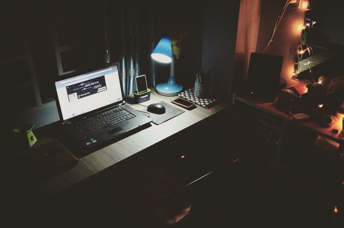 Home office desk at night with an open laptop, blue desk lamp, smartphone, and notebook on a wooden surface